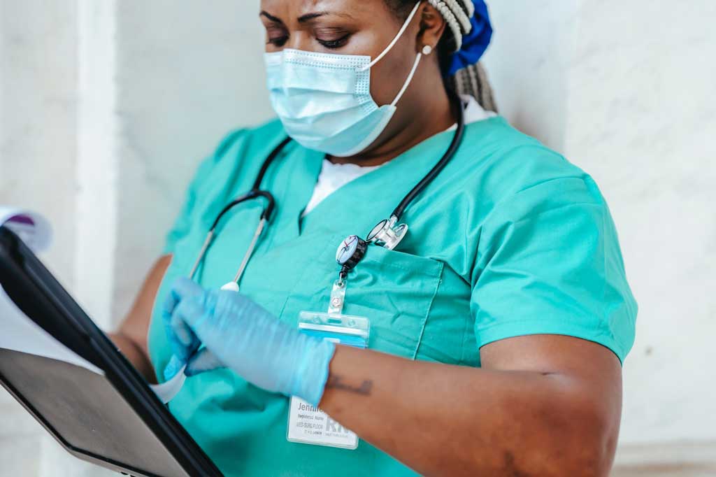 A nurse wearing masks and gloves looking at a clipboard.