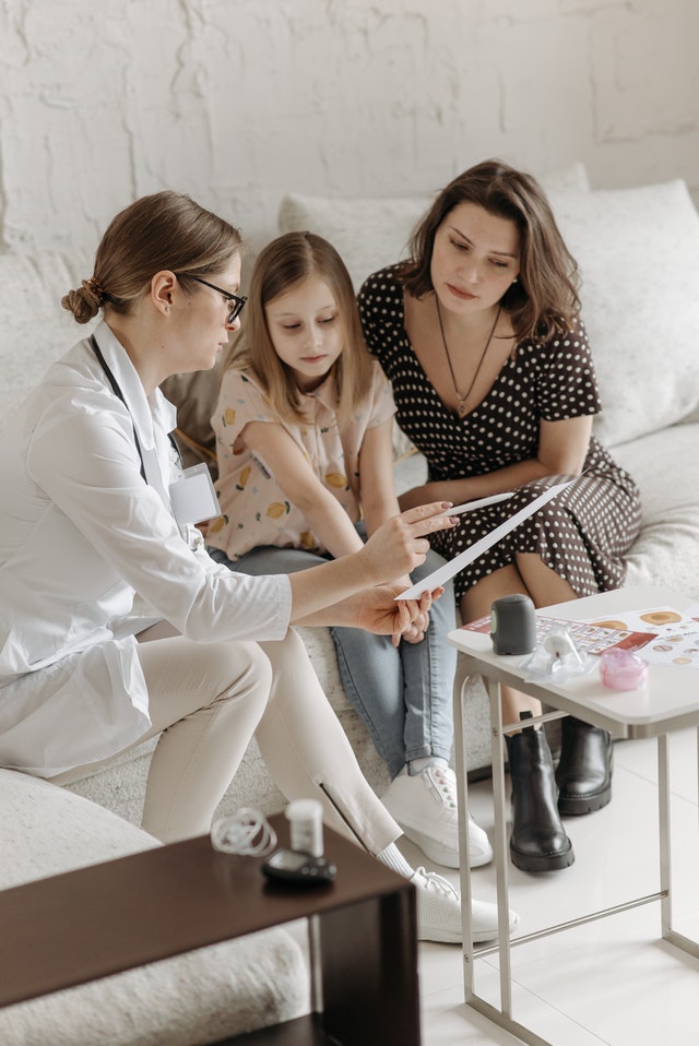 A doctor, child, and woman sitting on a couch looking at a paper that the doctor is pointing to