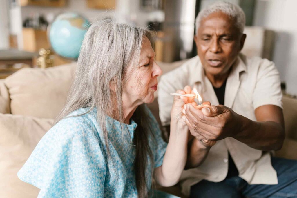 A heartwarming image of an elderly man assisting an elderly woman in lighting a joint, illustrating the practical use of Marijuana Strains and How They Can Help You manage age-related symptoms in a comfortable home setting.