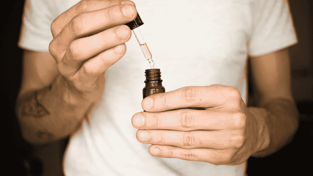 man in white t-shirt holding dropper and vial with yellowish liquid