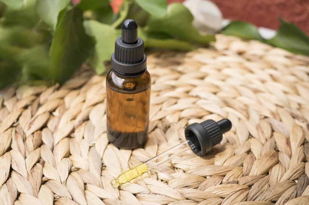 An amber glass bottle of CBD oil in a glass dropper vial resting on a woven straw mat, with the dropper lying beside it to show the golden, translucent liquid inside.