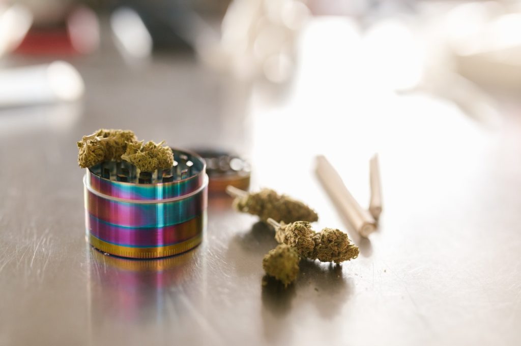 An artistic shot of Craft Cannabis buds sitting on an open iridescent, rainbow-colored metal grinder, with additional flower and a rolled joint resting in the soft, blurred background on a metal surface.