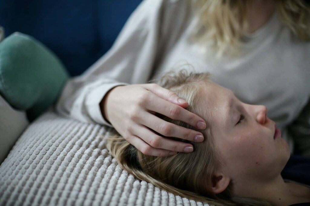 A tender, close-up shot of a mother’s hand gently resting on the forehead of her young daughter who is lying down with eyes closed, illustrating the parental care and medical consultation required when exploring How to Get a Medical Card Under 18.