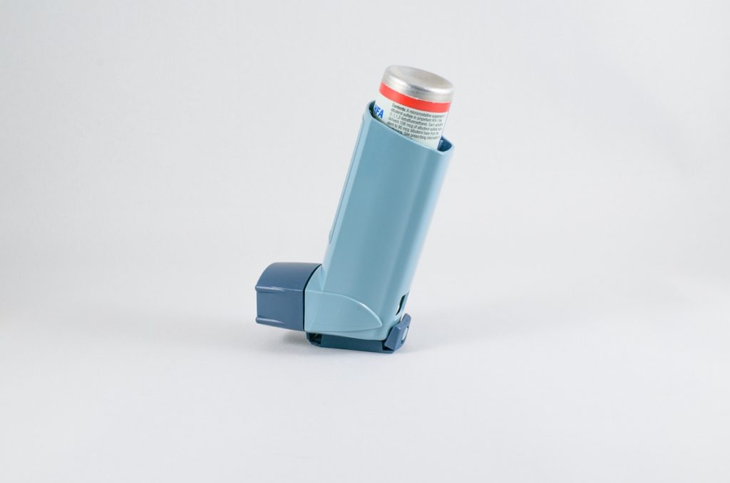 A side view of an inhaler device standing upright on a plain white surface, showing the light blue plastic casing and the metal canister containing respiratory medication.