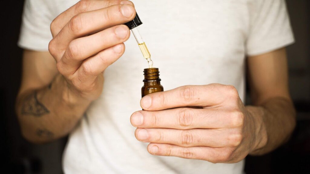 A close-up shot of a person’s hands holding an amber glass bottle and carefully lifting a glass dropper filled with golden THC-O Tincture against a white t-shirt background.