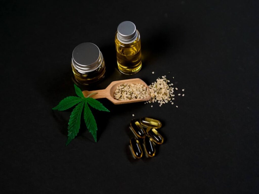An organized display of various Types of Cannabis Products, including two glass bottles of golden oil, several softgel capsules, a wooden scoop of hemp seeds, and a fresh green marijuana leaf, all set against a sleek black background.