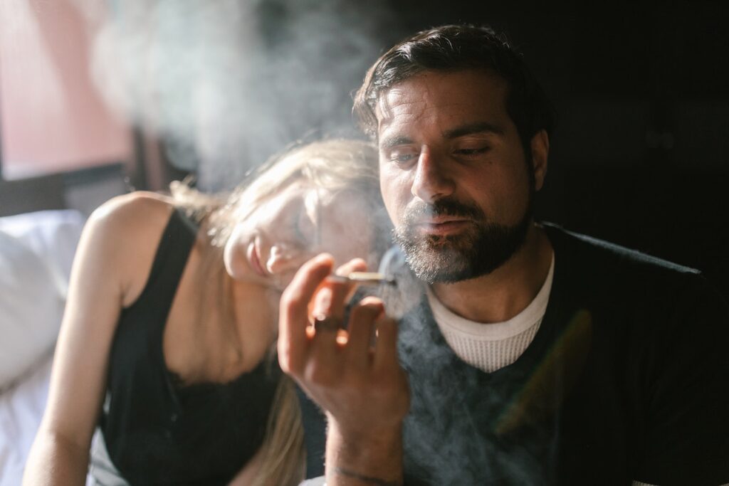 A moody, close-up shot of a man with a beard smoking marijuana, exhaling a thick cloud of smoke while holding a hand-rolled joint, with a woman leaning her head on his shoulder in the soft-focus background.