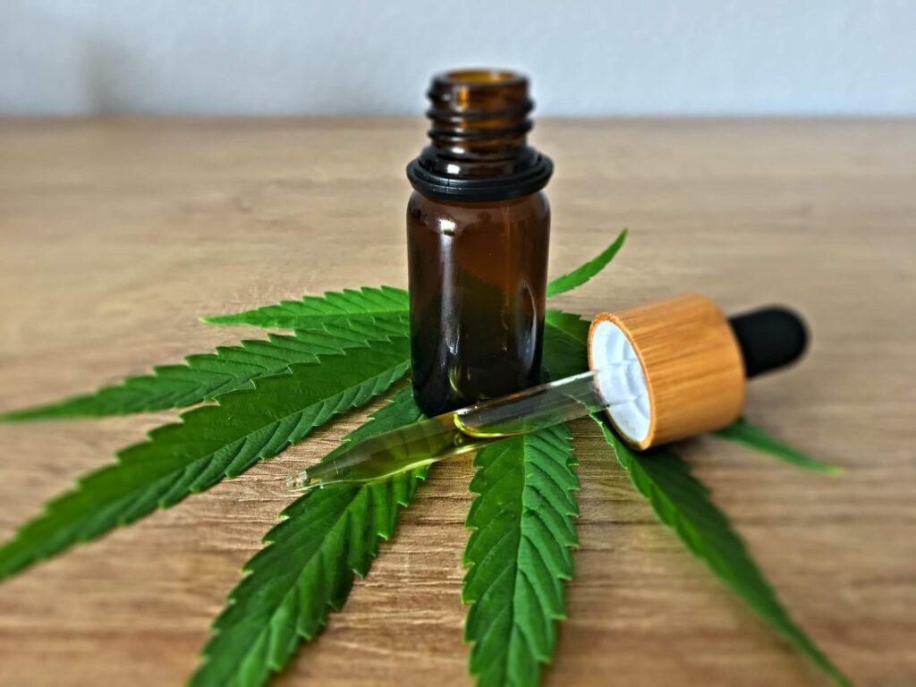 A small amber glass bottle and a glass dropper filled with golden Cannabis oil on a marijuana leaf, resting on a light wooden surface to showcase the natural botanical origins of cannabis-derived tinctures.