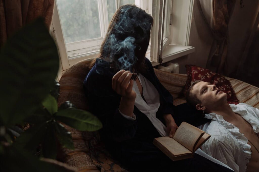 A cinematic, dimly lit scene of a couple smoking a blunt while relaxing on a vintage sofa near a window; one person holds an open book and exhales a swirl of smoke while the other rests their head in a moment of quiet contemplation.