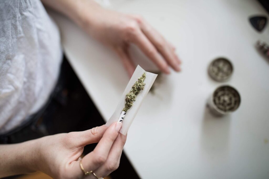 An overhead, close-up shot of a person’s hands carefully rolling a cannabis joint, holding ground medicinal flower inside a white rolling paper with a paper crutch filter, next to a metal herb grinder on a white table.