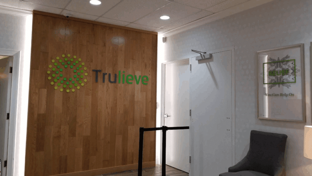 Trulieve dispensary lobby interior featuring the brand logo on a vertical wood-paneled wall, a stanchion barrier, and a waiting area with a grey armchair.