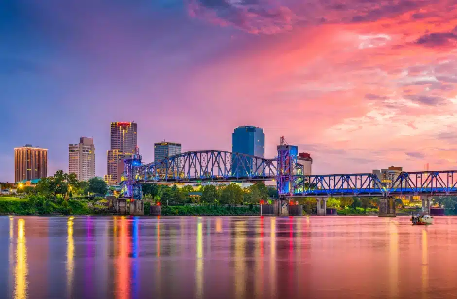 A beautiful twilight skyline view of Little Rock, Arkansas, featuring illuminated city skyscrapers and a steel truss bridge stretching across the Arkansas River, with vibrant pink, purple, and orange sunset clouds reflecting on the calm water surface.