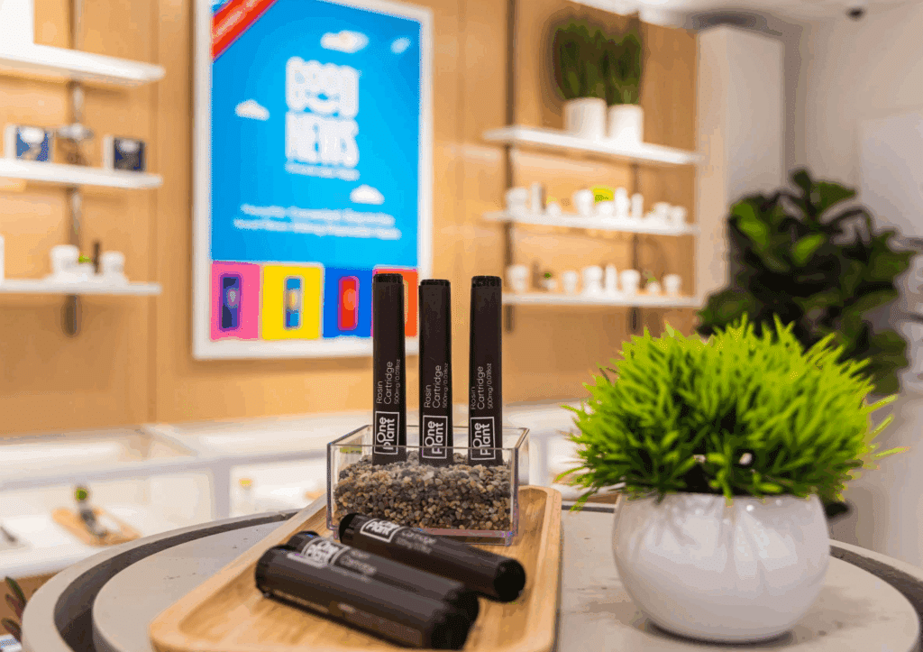 One Plant Rosin Cartridges displayed on a retail countertop inside a bright dispensary, with a vibrant "Good News" brand poster visible in the blurred background.