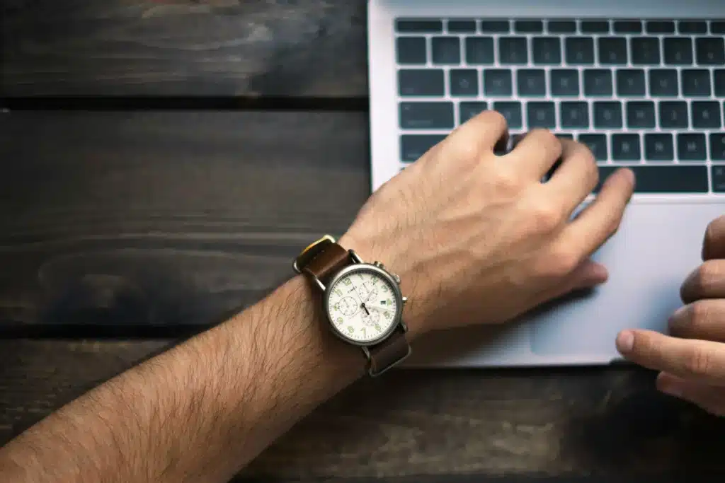 Person using a laptop while checking time on a wristwatch, representing how long does it take to get an MMJ card in Pennsylvania.