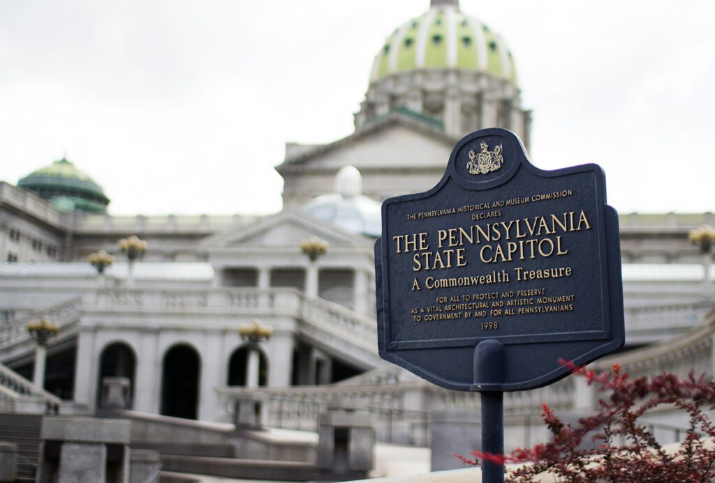 A close-up photograph of a blue and gold historical marker reading "The Pennsylvania State Capitol: A Commonwealth Treasure," with the blurred dome and grand architecture of the Capitol building in Harrisburg standing in the background.