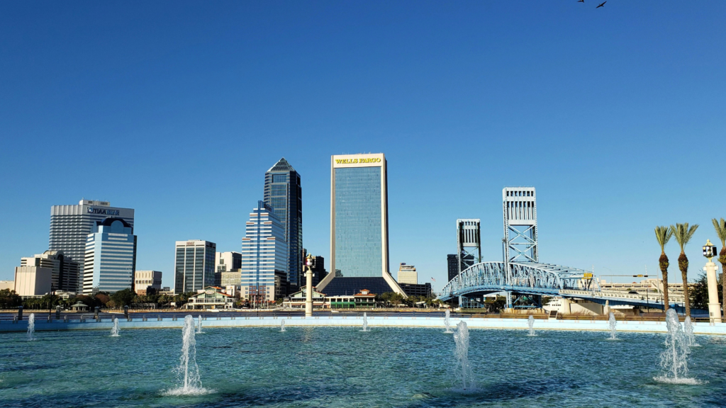 A sunny, wide-angle view of the Tampa city skyline with skyscrapers reflecting across the water and palm trees on the side.