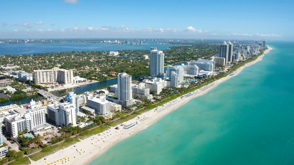 A high-angle aerial view of a coastal city shoreline, showing white high-rise buildings running alongside a sandy beach and turquoise ocean water.