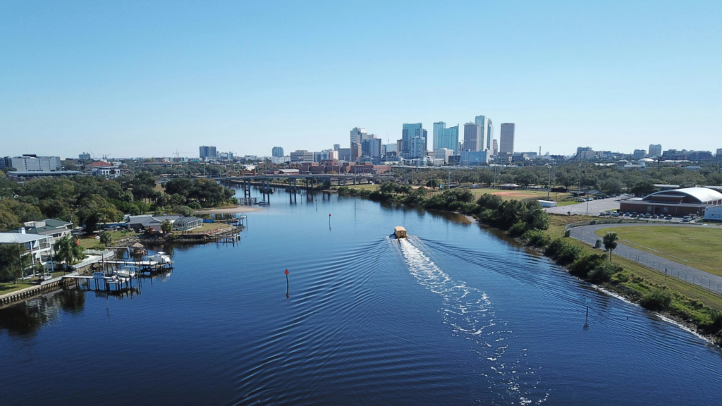An aerial view of a wide, blue river cutting through a city landscape under a clear sky, with a boat leaving a wake in the water.