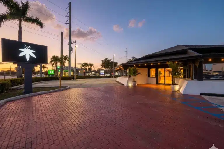 Exterior view of The Flowery Palmetto Bay, a medical marijuana dispensary in Florida, featuring its signature flower logo sign and red brick driveway.